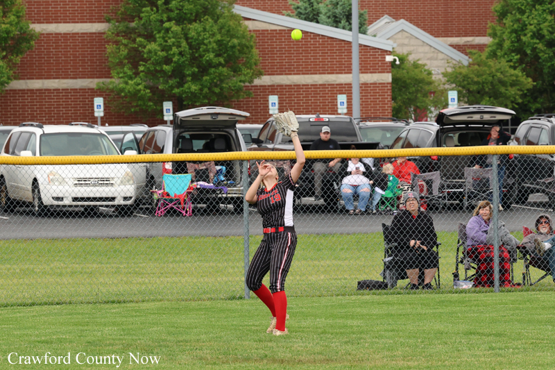 Softball player in a black pinstripe uniform reaches up to catch a fly ball near the outfield fence as spectators sit in chairs behind the chain-link fence.