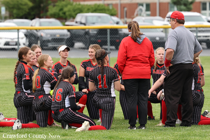 Girls softball team kneeling in a huddle on the field, listening to two coaches (one in red jacket, one in gray) at practice.