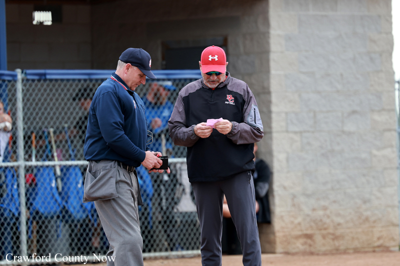 Two men in baseball attire stand near a dugout, one in a navy cap examining a small device while the other reads a pink note.