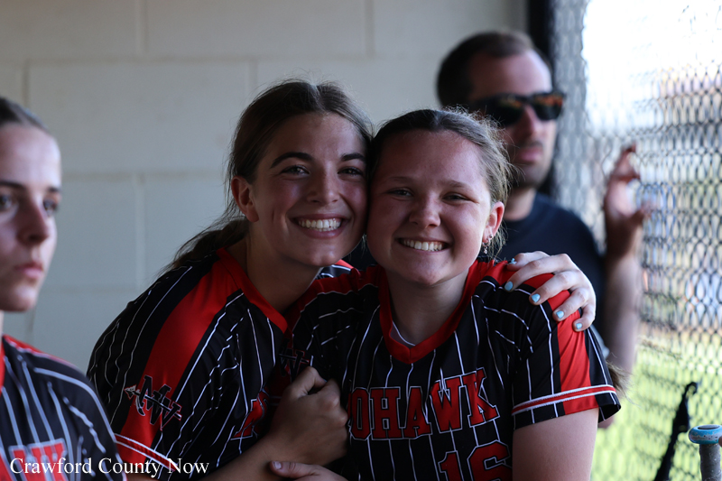Two smiling female softball players in black-and-red striped jerseys hug in a dugout, with a man in sunglasses standing behind them near a chain-link fence.