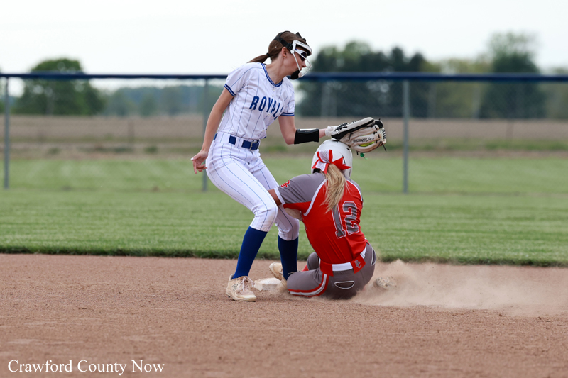 Softball play at a base: a white-and-blue-uniform runner stands by the bag as a red-uniform fielder slides into the base, kicking up dust.