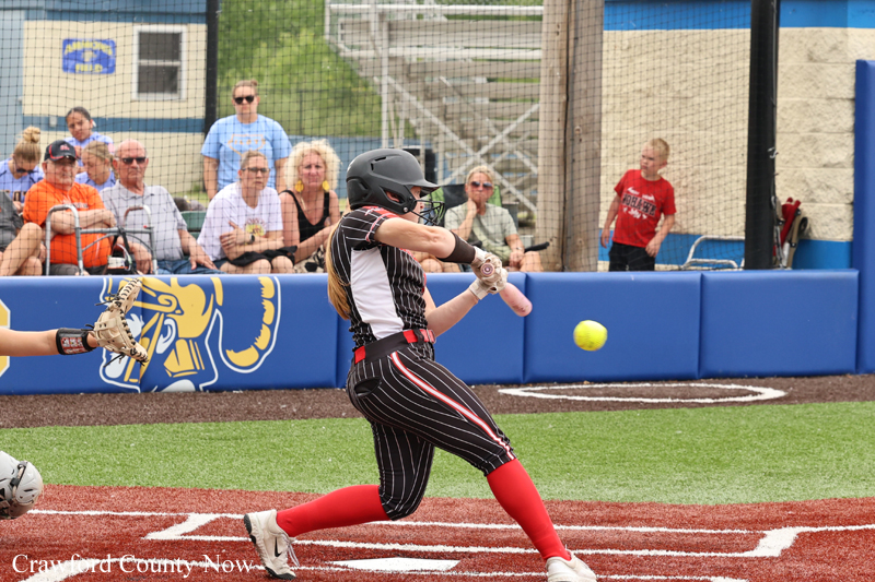 Female softball batter in a black-and-white pinstripe uniform swings at a pitched ball during a game, with spectators watching from behind the dugout fencing.
