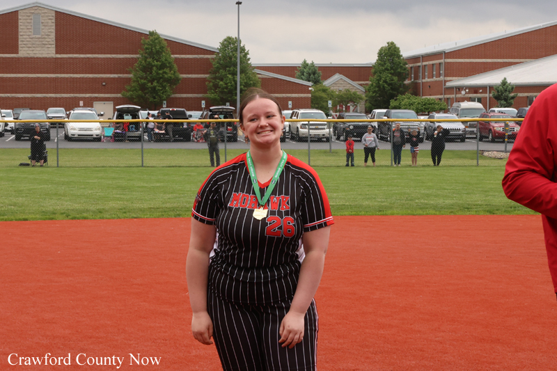 Softball player in pinstripe uniform with number 26 and medal, standing on a red dirt infield with spectators and school buildings in the background.