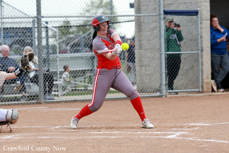 Softball batter in red and gray swings at a pitch on a dirt field, catcher and spectators behind a chain-link fence in the background.