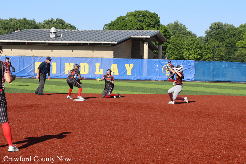 Softball game on a sunny day: batter swings at home plate while four fielders in maroon uniforms position around the infield with a blue outfield wall behind them.