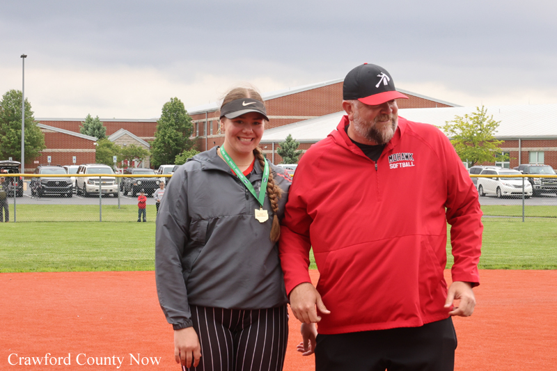 Smiling young female softball player with medal around her neck walks beside a bearded man in a red jacket on a softball field; school buildings in background.