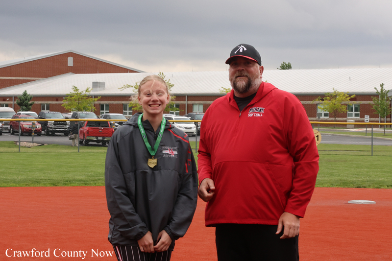 Young female softball player wearing a green medal ribbon stands with a man in a red jacket on a softball field, with a school building in the background.