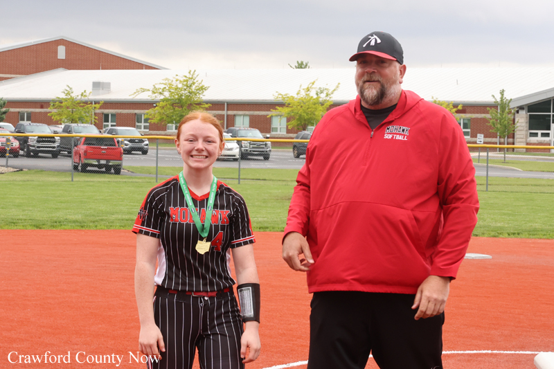 Young girl in a black pinstripe softball uniform with a gold medal, standing on the orange infield beside a man in a red jacket on a softball field.