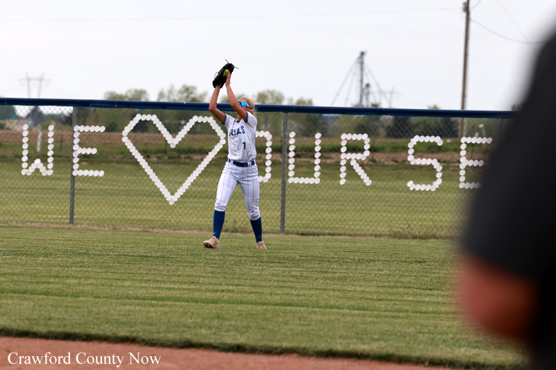 Softball player in white uniform catches a ball in the outfield, with a fence banner that reads 'WE ♥ OUR SE' in the background.