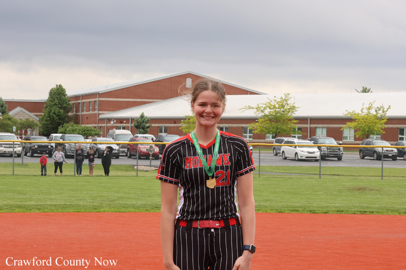 Young female athlete in a pinstripe uniform with gold medals around her neck, smiling on a sports field with a red infield behind her.