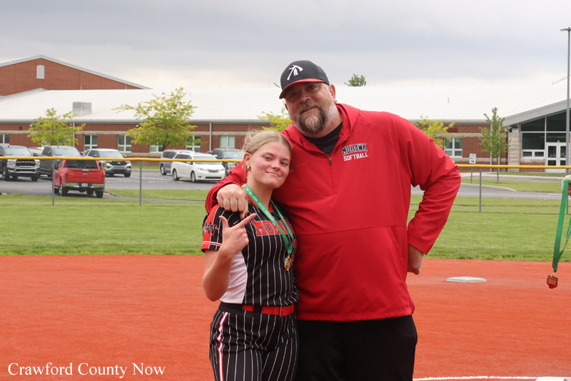Young softball player with medals posing with a coach in a red jacket on a softball diamond.