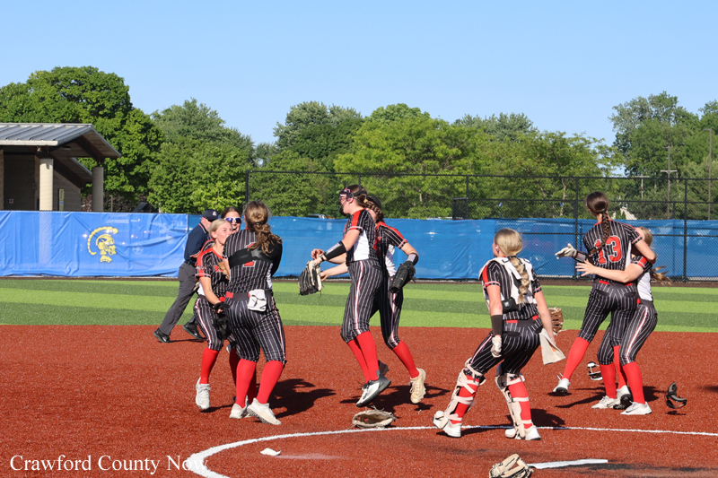 Softball players in black pinstripe uniforms celebrate on the red infield, red socks visible, gloves in hand on a sunny day.