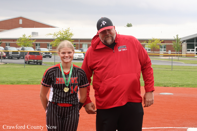 Girl in a black pinstripe softball uniform with a medal around her neck poses with an adult coach in a red jacket on an orange softball infield after a game.
