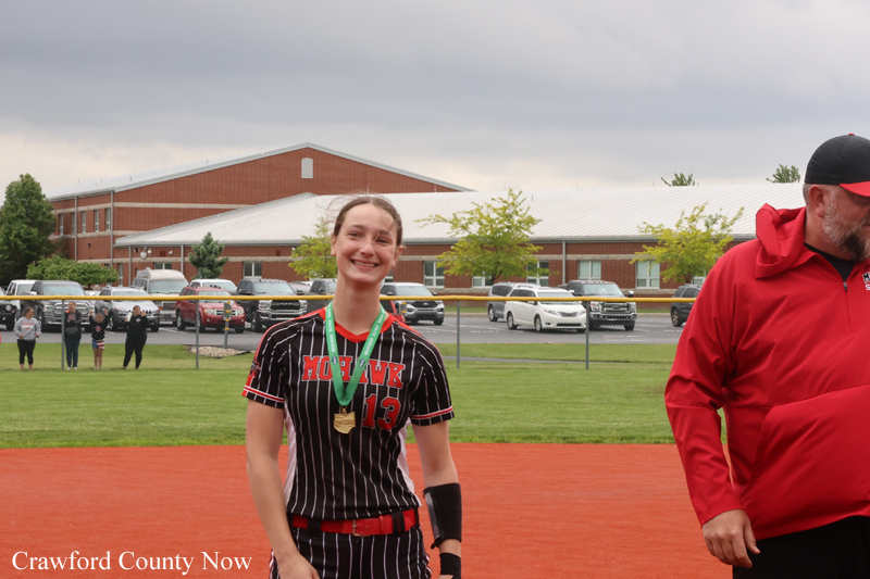 Young female softball player in a black pinstripe uniform with number 13, medal around her neck, smiling on a baseball/softball field while a coach in a red jacket stands nearby.
