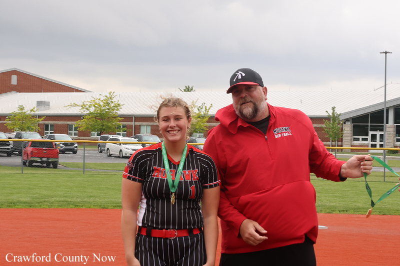 Young female softball player with a medal posing beside her coach on a softball field.