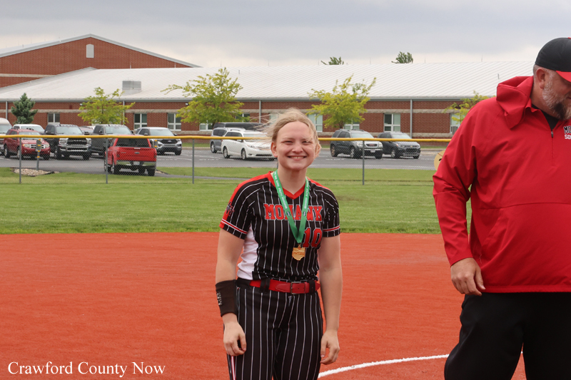 Young girl in a black pinstripe softball uniform with a gold medal on a green ribbon, posing on a red infield with a man in a red jacket nearby.