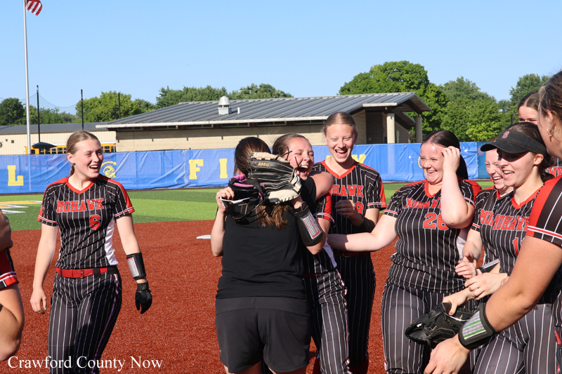 Group of girls in black pinstripe softball uniforms celebrate together on the infield, smiling and hugging near a glove-wielding teammate on a sunny day.