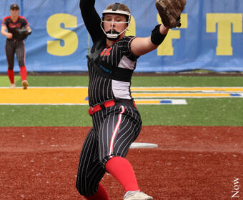 Softball pitcher in a black pinstripe uniform mid-throw on a red infield, yellow ball raised in her right hand with glove ready on her left hand.