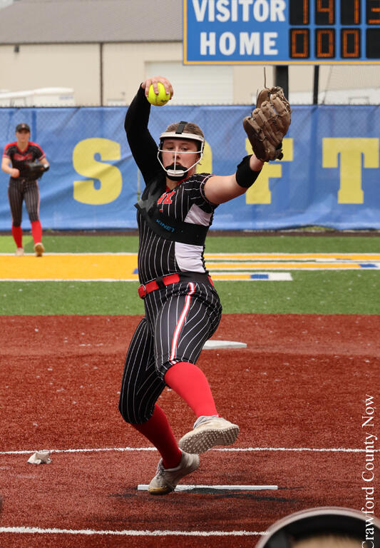 Softball pitcher in a black pinstripe uniform mid-throw on a red infield, yellow ball raised in her right hand with glove ready on her left hand.