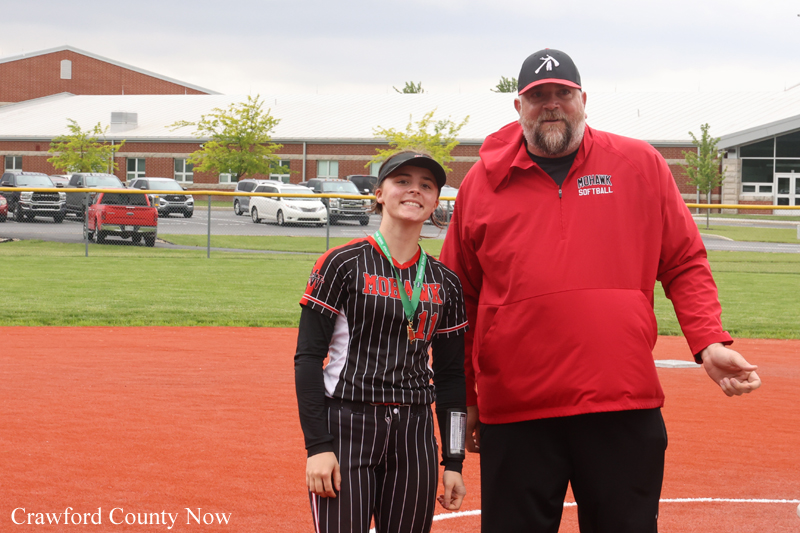Young female softball player in a pinstriped uniform with medal posing with a coach on an orange-dirt softball field near a school.