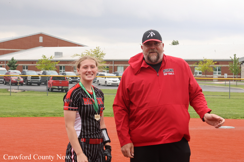 Young female softball player with medals posing with a coach in a red jacket on a softball field after a game.