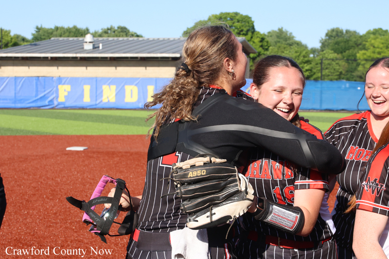 Three female softball players in black and red uniforms hug and smile on the field after a game.