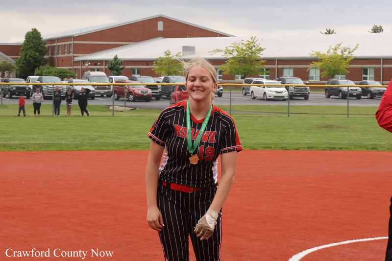 Smiling female softball player in a pinstripe black and red uniform with a gold medal, standing on a red-dirt field after a game.