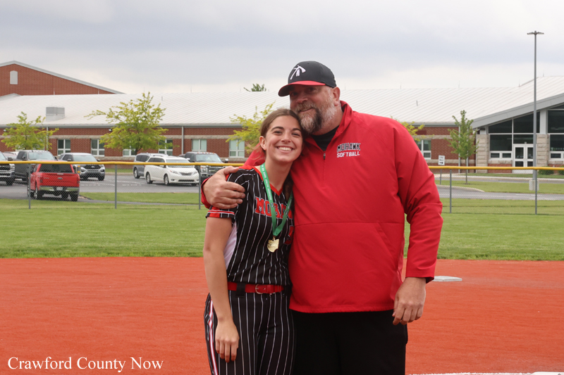 Young female softball player in pinstriped uniform with a medal, posing and smiling with a coach on a red clay field outside a school campus.