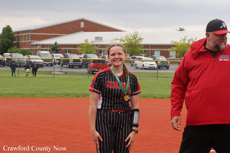 Smiling young softball player in a pinstriped uniform with a medal around her neck on a field, with a coach in a red jacket nearby.