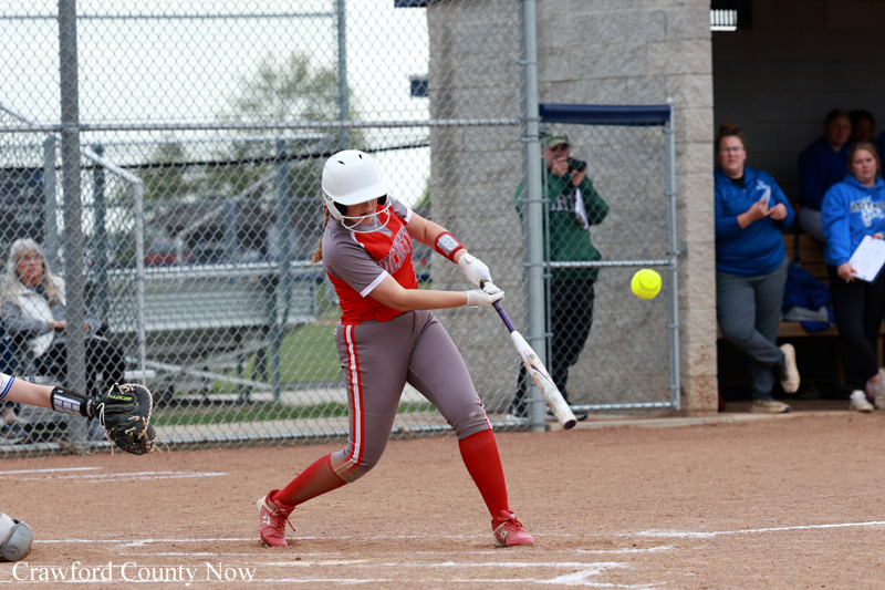 Softball batter in red swings at a pitched ball during a game, with catcher and dugout in background behind chain-link fence.