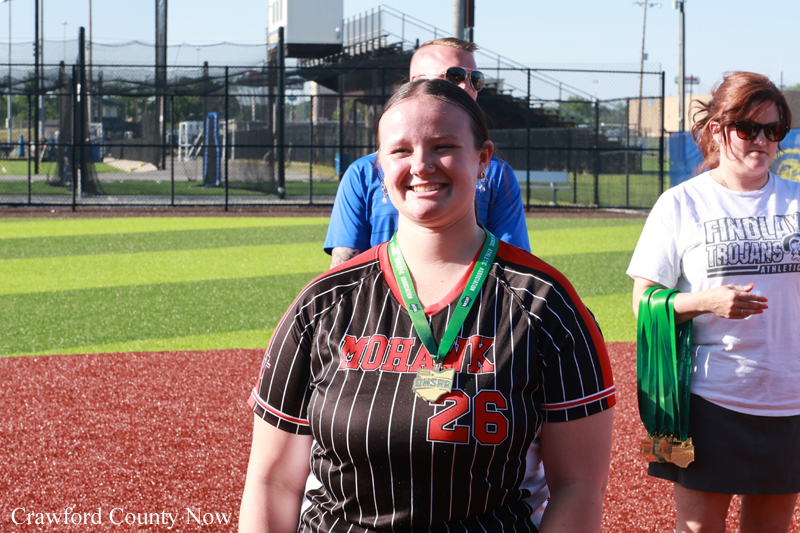 Smiling woman in a black pinstripe Mohawk jersey (#26) with a gold medal on a green ribbon, on a sports field with teammates behind her.