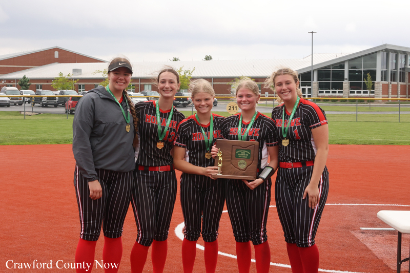 Softball team posing on the field with medals and a plaque trophy after a win, five players and coach smiling.