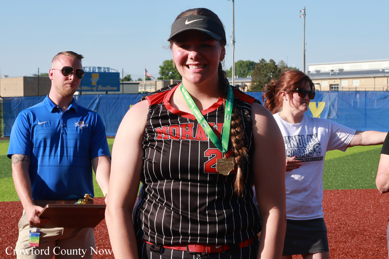 Smiling female softball player in a pinstriped jersey with a gold medal around her neck, standing on a sports field with two people behind her.