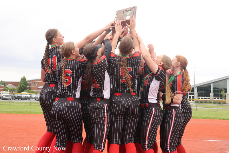 Girls softball team in black pinstripe uniforms huddle and lift a trophy in celebration on a dirt field infield edge at a sports complex.