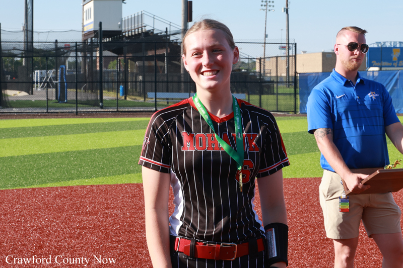 Smiling young female athlete with a medal on a green lanyard, standing on a sports field; a man with a clipboard is in the background.
