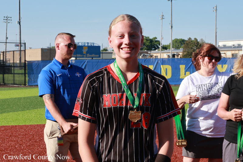 Young female athlete wearing a black pinstripe jersey with red lettering and a green medal, smiling at a sports awards event on the field.