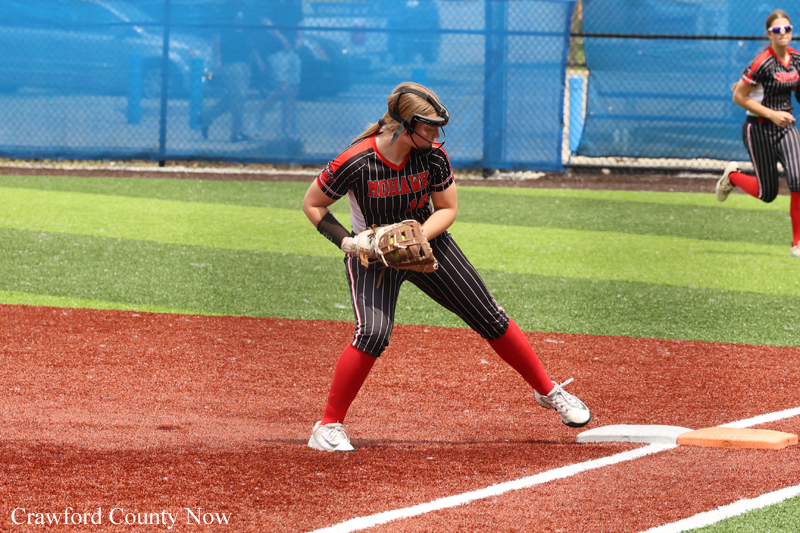 Female softball player in black pinstripe uniform and red socks catching a ground ball near first base on a turf field.