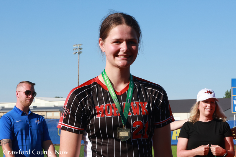 Young female athlete with a gold medal on a green ribbon, wearing a black pinstripe jersey, smiling on a sports field.