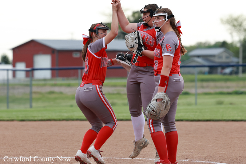 Four female softball players in red uniforms high-five on the dirt infield on a sunny day in the field.
