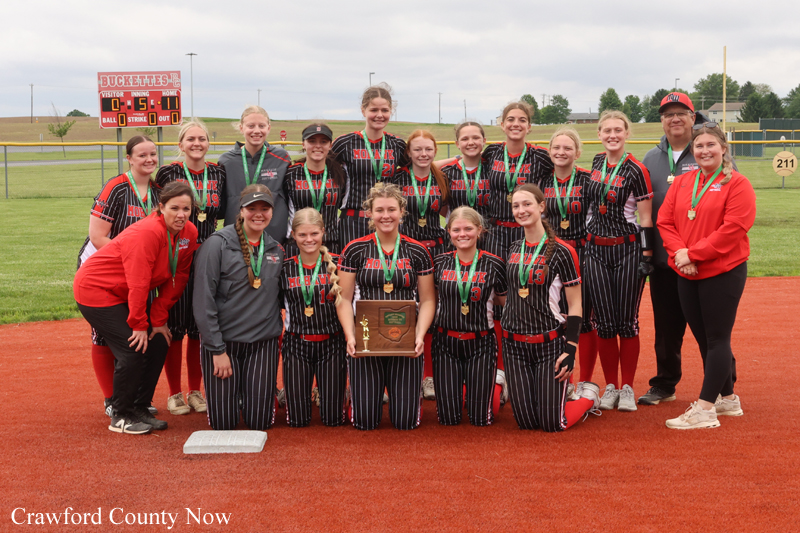 Girls' softball team in black pinstriped uniforms with red trim posing on a baseball diamond, holding a trophy while wearing medals; coaches stand at the sides.