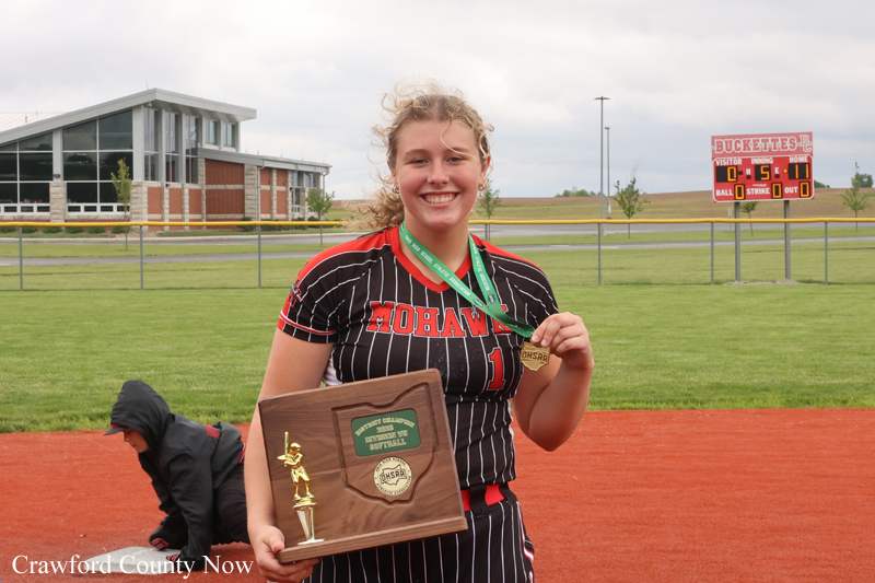Softball player in a black pinstriped Mohawks uniform smiles, holding a wooden trophy and green medal on a grassy field with a scoreboard in the background.