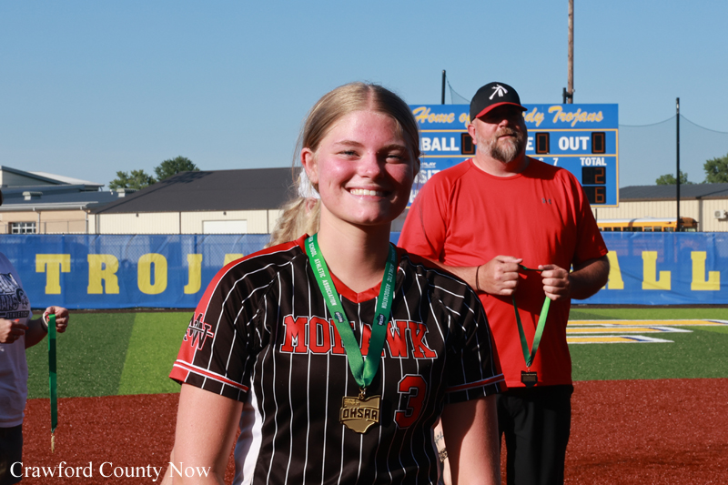 Smiling young female softball player in a black and red striped uniform with a gold medal on a green ribbon, on a baseball field with a scoreboard in the background.
