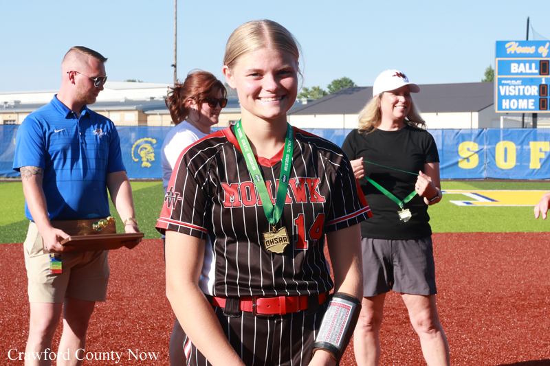 Smiling female softball player in a black pinstripe uniform with a medal on a red dirt field; award ceremony in the background.
