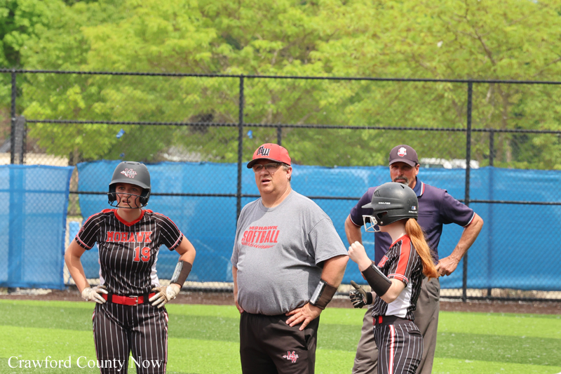 Softball players and coaches stand on a sunny field behind a chain-link fence, wearing helmets and uniforms.