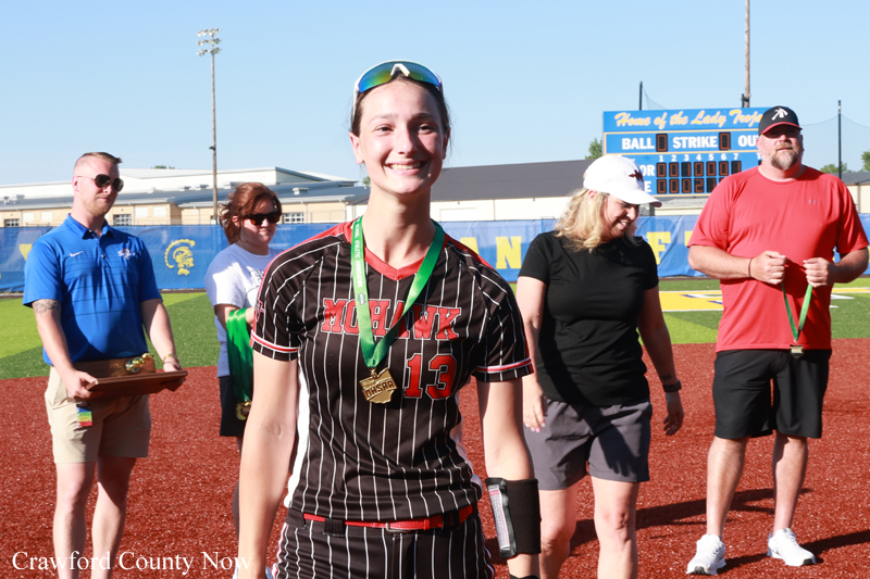 Smiling female softball player with a gold medal on a green lanyard standing on a softball field during an award ceremony, with teammates in the background.