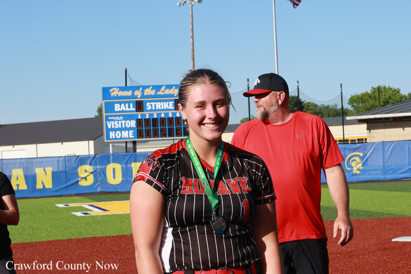 Smiling softball player in a black pinstripe uniform with red lettering and a green medal around her neck, on a baseball field with a scoreboard in the background.