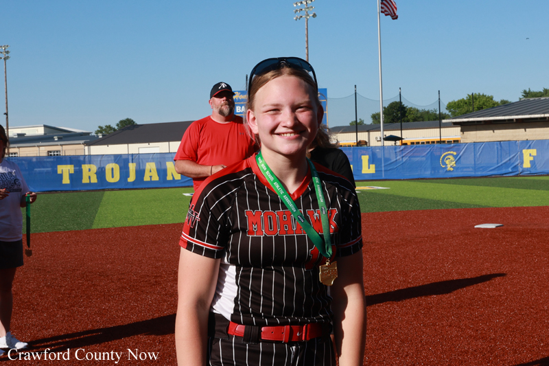 Smiling young softball player in a black pinstripe uniform with a gold medal on a green ribbon stands on a red infield; blue outfield wall behind her reads 'TROJANS'.
