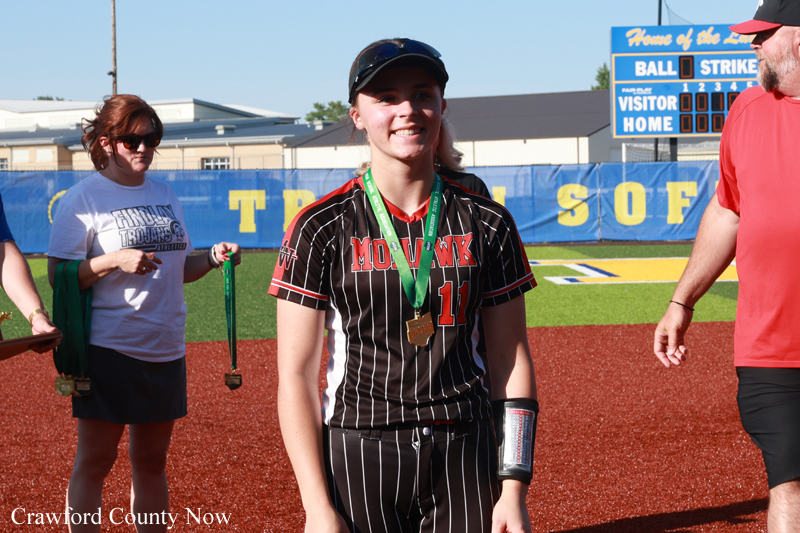 Smiling young softball player in a black pinstripe uniform with medals around her neck, standing on the field with teammates and a scoreboard in the background.