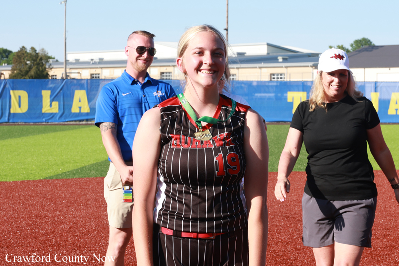 Smiling female track athlete wearing number 19 with a medal, standing on a red track with two supporters behind her.