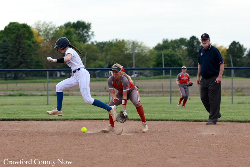 Softball action: a runner in white and blue slides toward a base as a fielder in red gloves the ball near the dirt. An umpire watches from the right field.